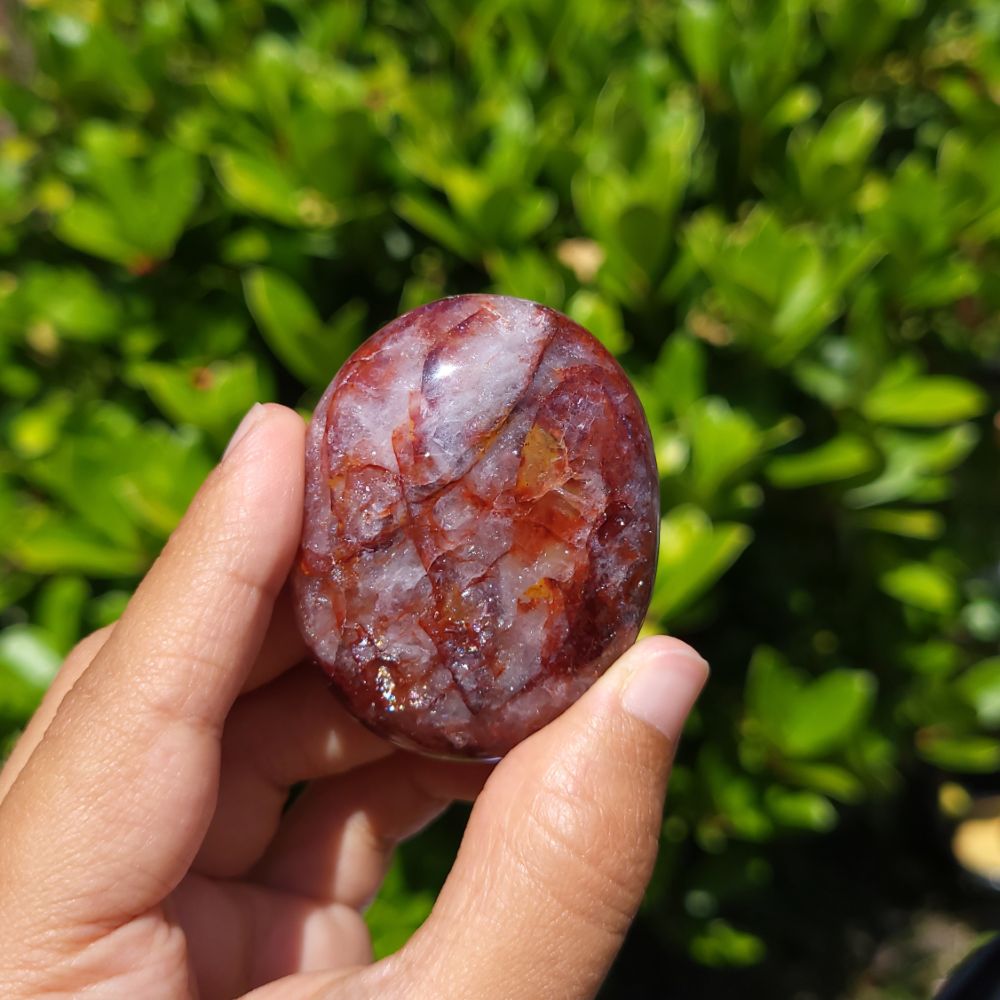 Hand holding a polished fire quartz palm stone against a green leafy background