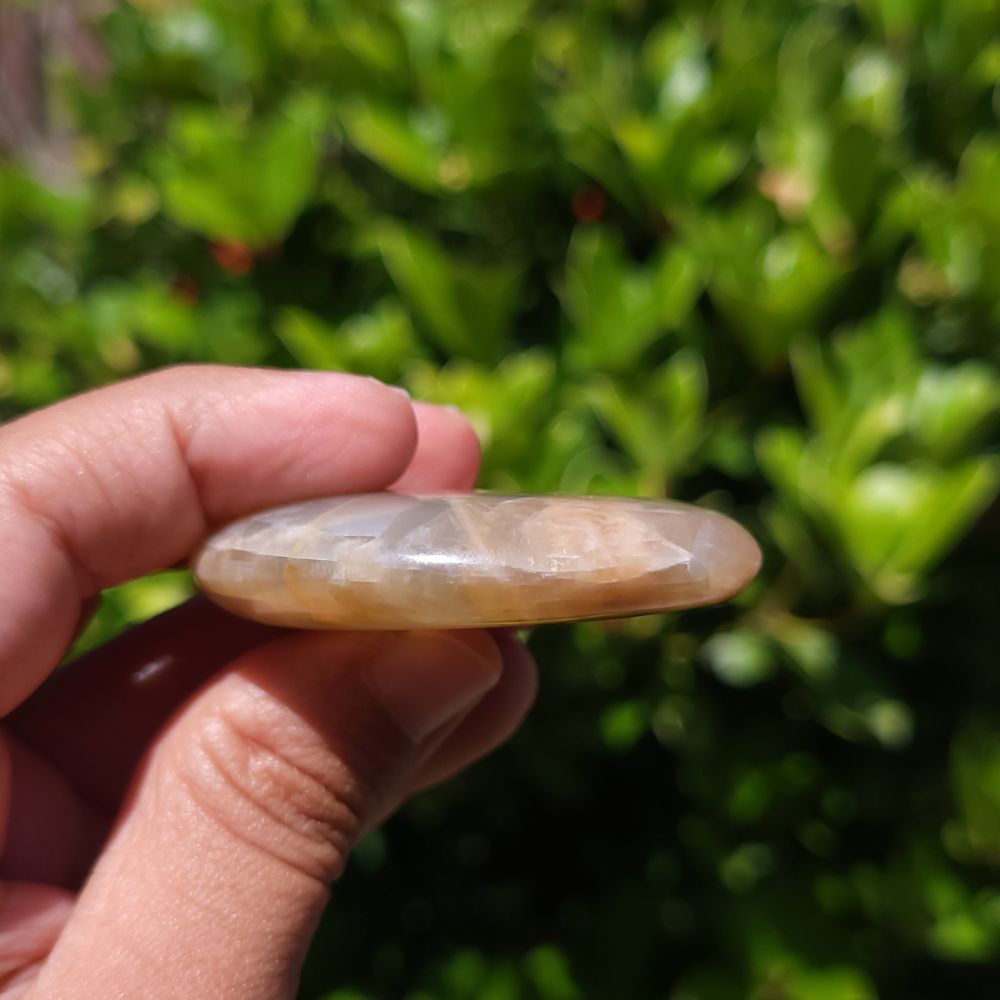Hand holding a smooth, round peach moonstone palm stone against a blurred green background