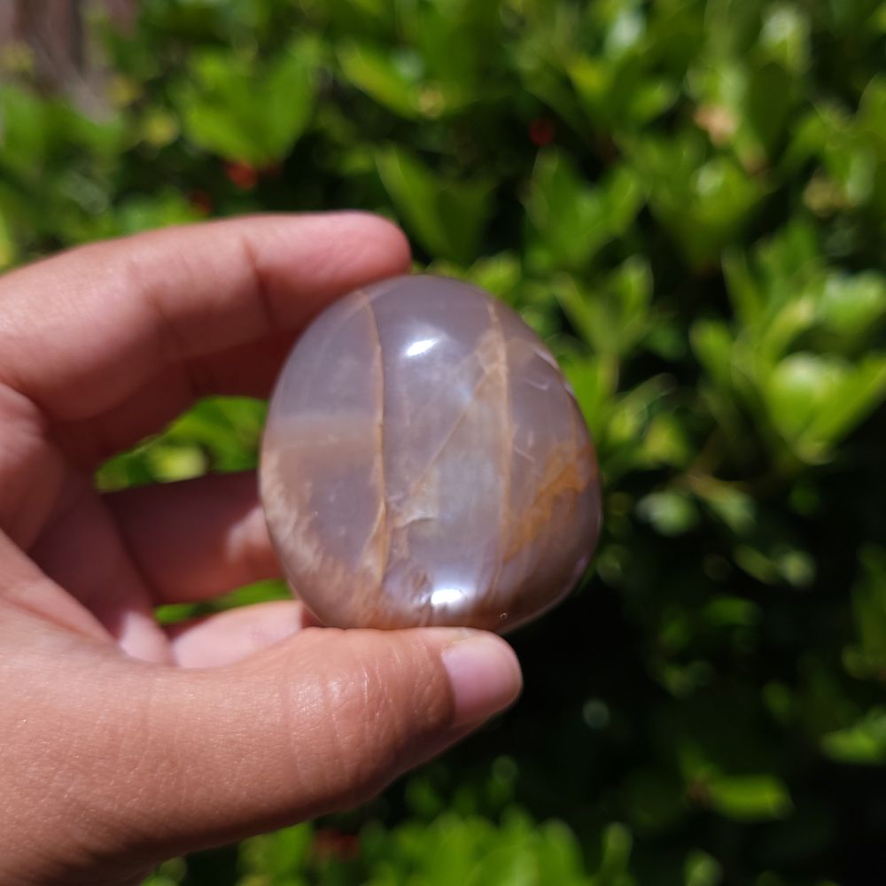 Hand holding a polished peach moonstone palm stone with a natural pattern against a blurred green background