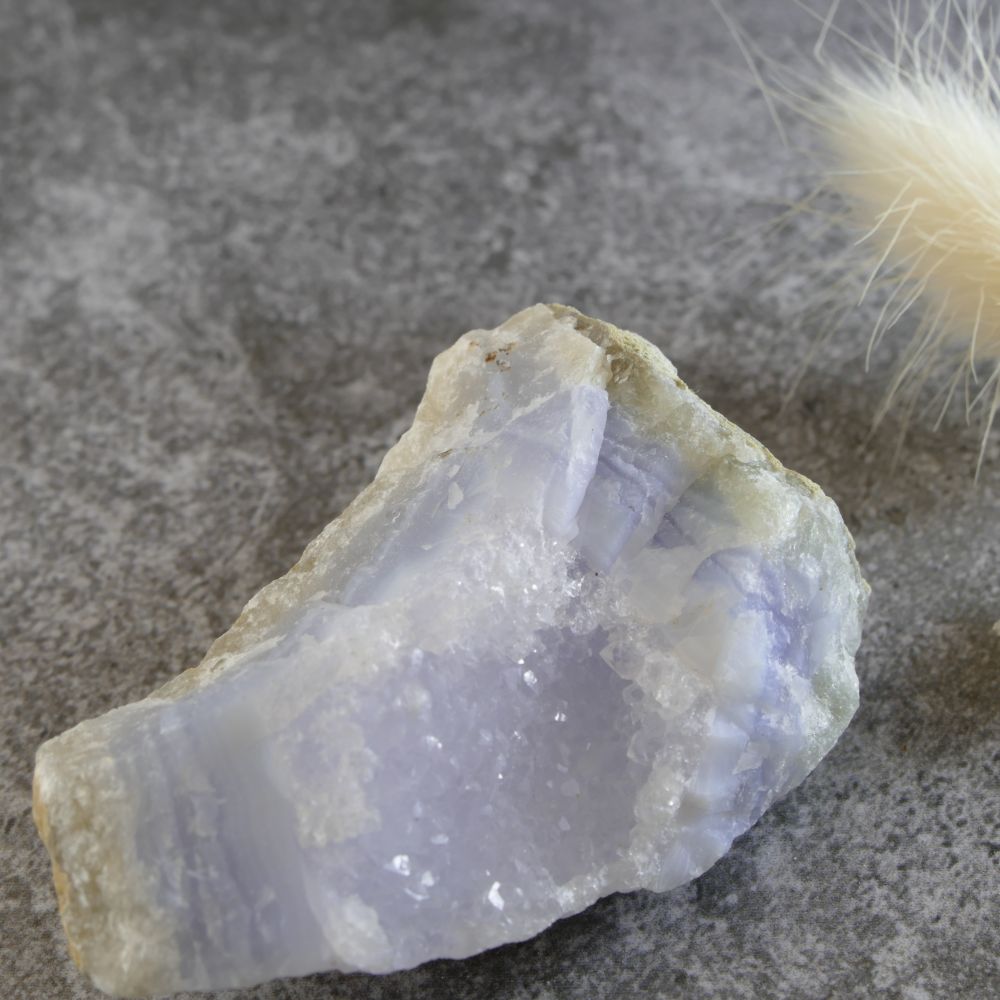Close-up of a raw, unpolished blue lace agate specimen with soft blue and white banding, featuring naturally formed, sparkling quartz crystals. Highlights the texture and unique crystal formations.