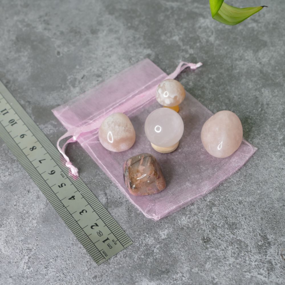 Set of pink amethyst, rhodonite, rose quartz tumbled stones, flower agate mushroom and rose quartz mini sphere on a pink drawstring bag with a ruler for scale on a gray surface.