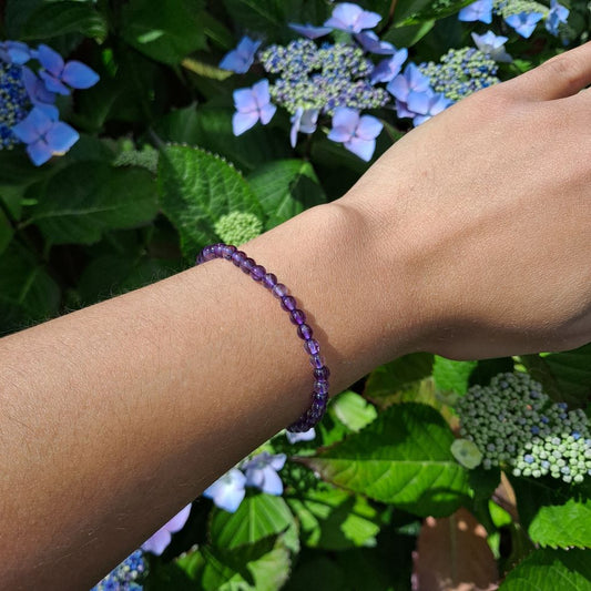 A stretch amethyst handmade bracelet with 4mm round beads, elegantly worn on a wrist, from Dumi's Crystals. The deep purple hues of the amethyst beads are highlighted against the skin, showcasing the bracelet's delicate and sophisticated design.