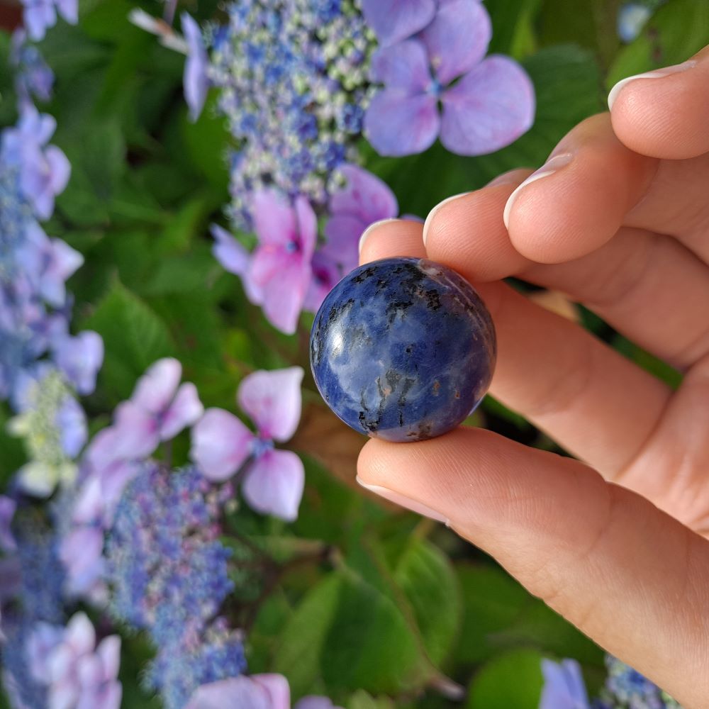 Dumi's Crystals | Sodalite Sphere (25mm) | A close-up view of a captivating Sodalite Sphere (25mm), showcasing its vibrant blue hues with flecks of white. This substantial stone, known as the Stone of Logic, is believed to promote mental clarity, peace, and clear communication. It's also thought to reduce stress and boost confidence.