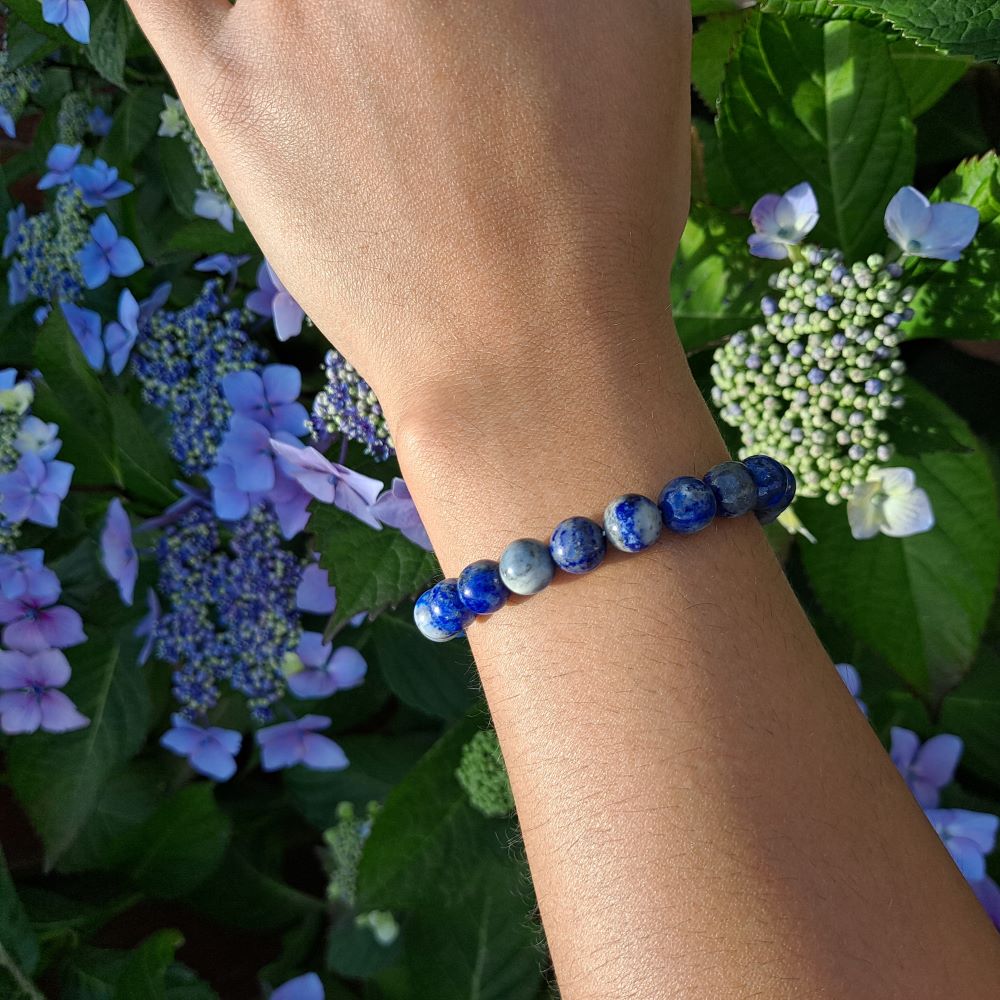 hand showing a blue lapis lazuli beaded bracelet, green leaves and purple flowers in the background