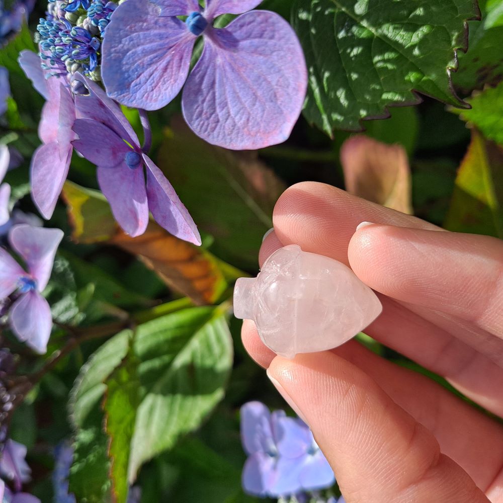 pink rose quartz hazelnut carving held in a hand, green leaves and purple flowers in the background