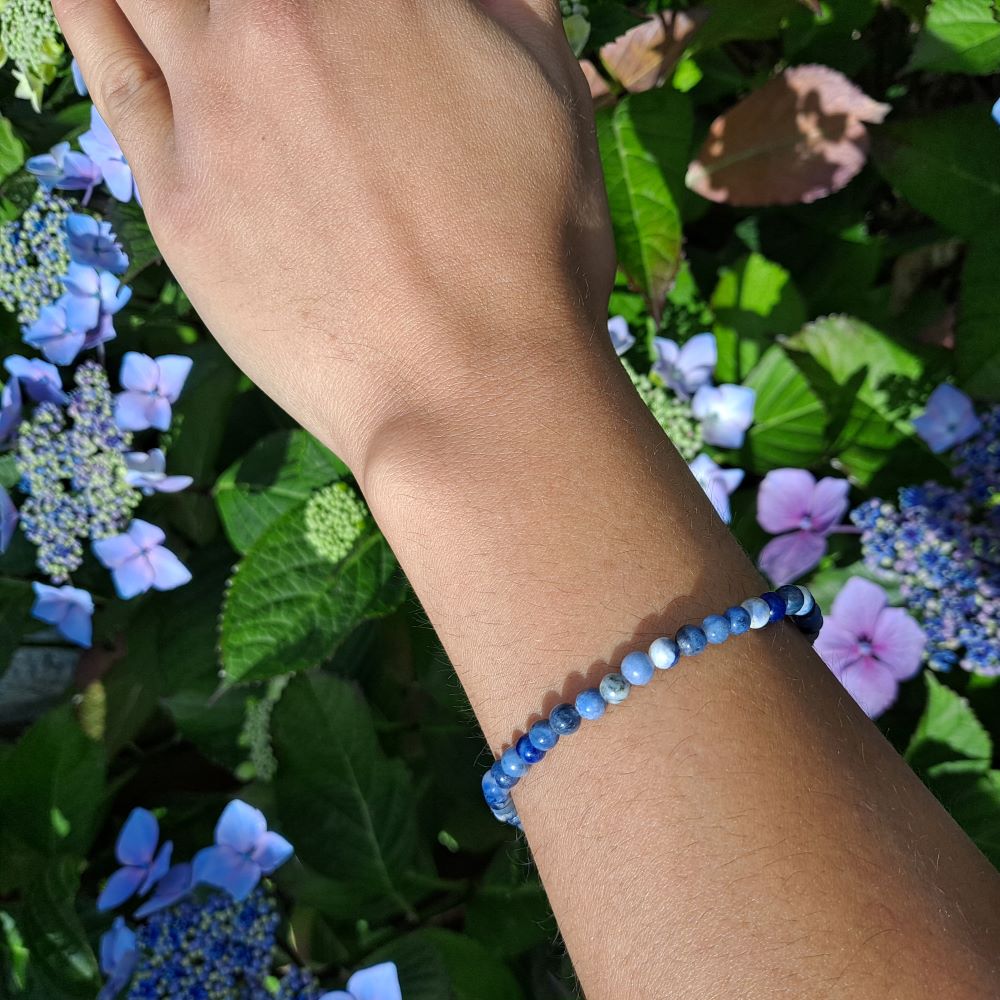 hand showing a blue sodalite beaded bracelet, green leaves and purple flowers in the background