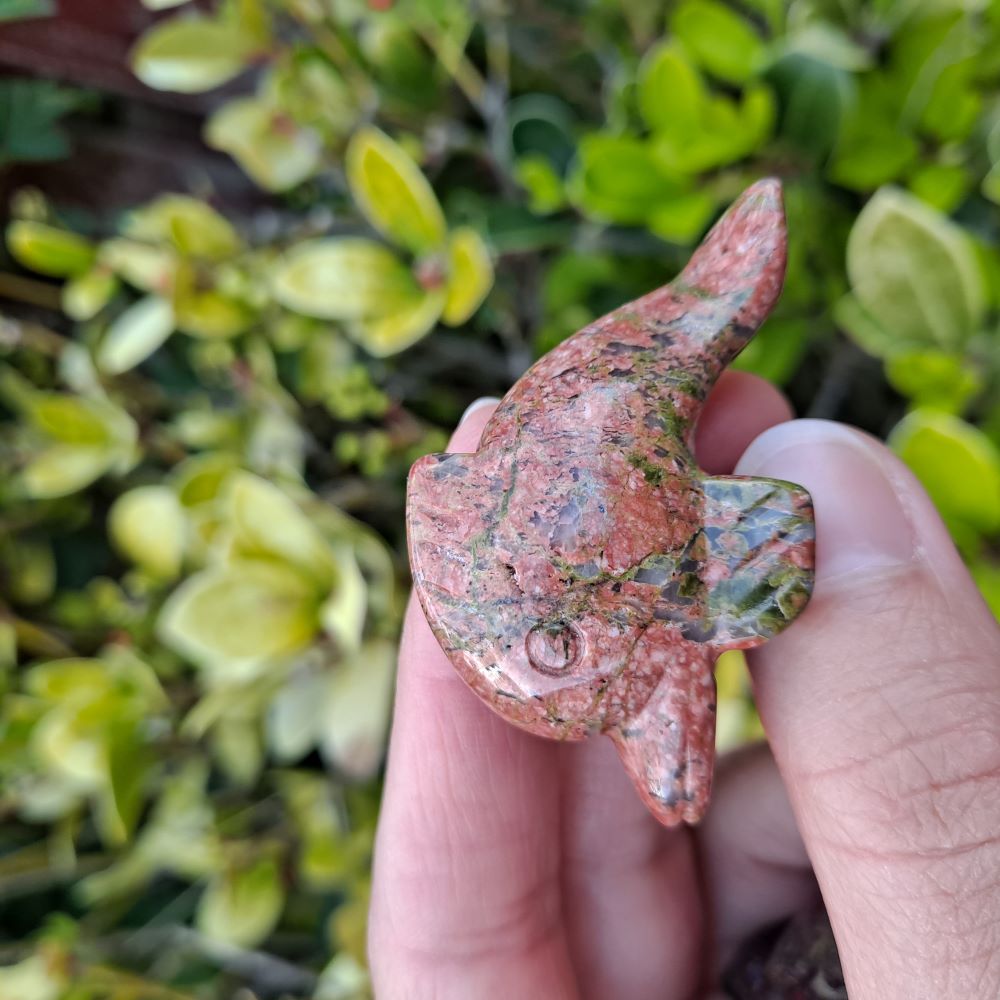 green and pink unakite crystal dolphin carving held in a hand, green leaves in the background