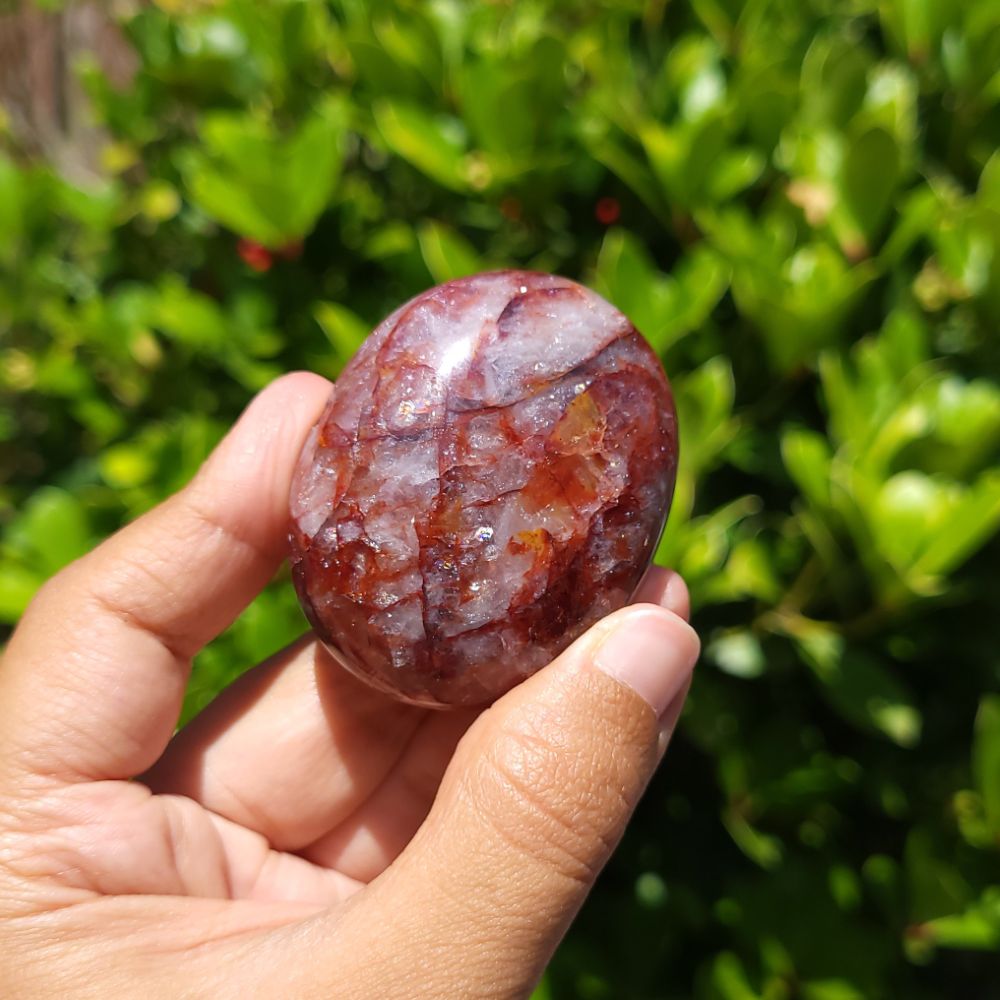 Hand holding a polished fire quartz palm stone with a natural marbled pattern against a green leafy background