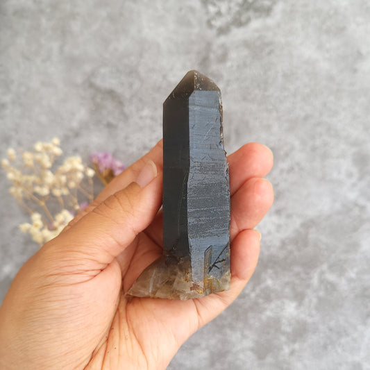 A hand holding a dark gray smoky quartz crystal point against a textured gray background with small dried flowers in the corner.
