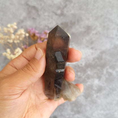 A hand holding a dark brown smoky quartz crystal point against a textured gray background with blurred dried flowers.