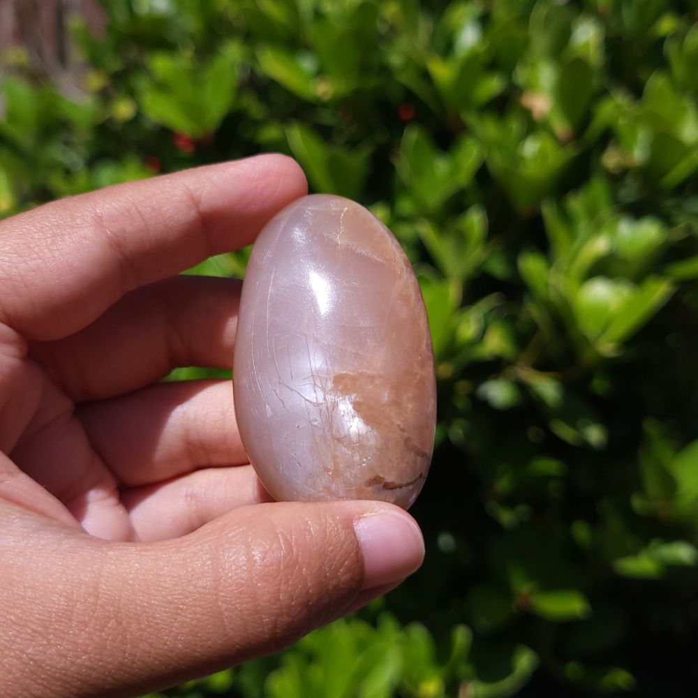 Hand holding a peach moonstone palm stone with a green outdoor background