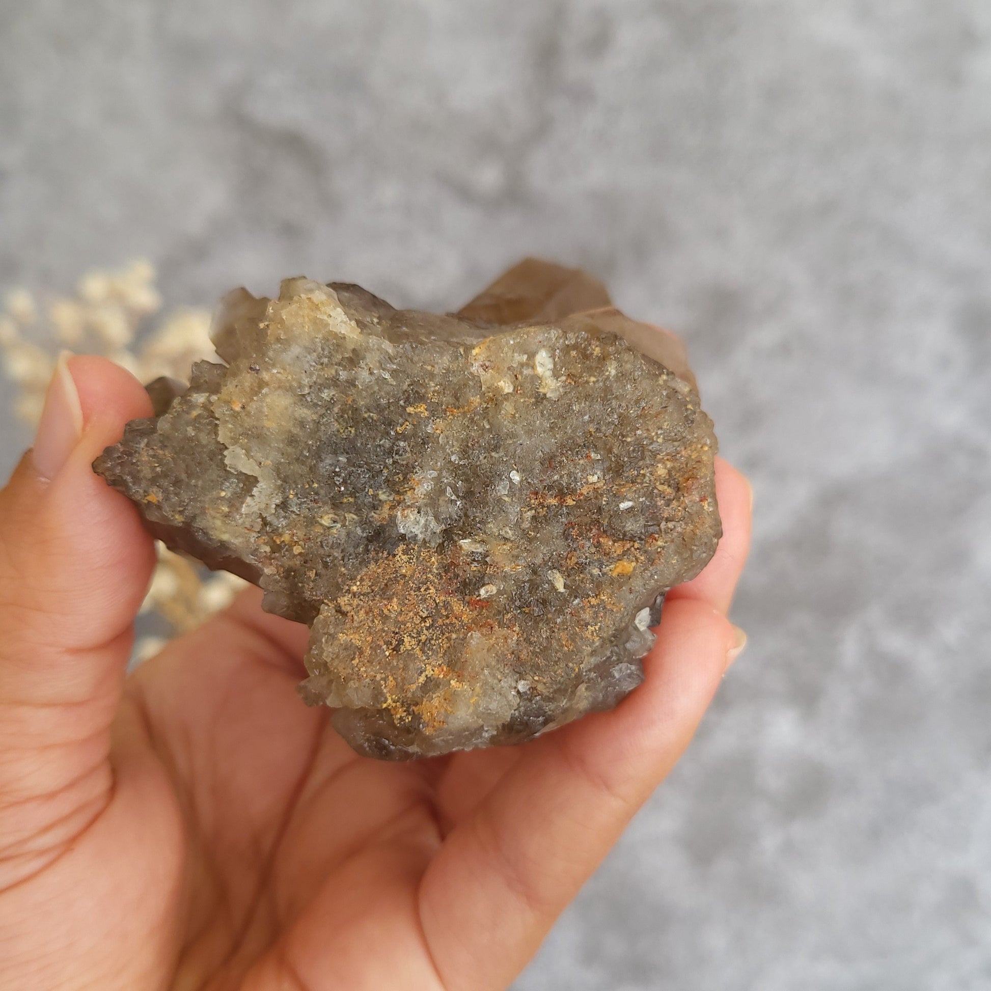 A hand holding a rough, natural mineral or crystal specimen with a mix of gray, brown, and metallic gold tones against a blurred gray background.