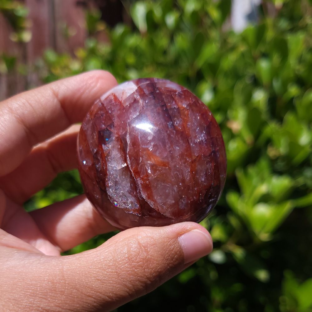 Hand holding a Hematoid Quartz palm stone with a blurred green outdoor background