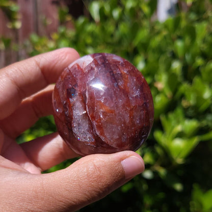 Hand holding a Hematoid Quartz palm stone with a blurred green outdoor background