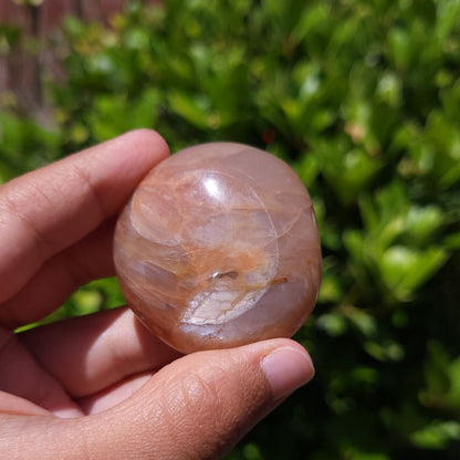 Hand holding a polished peach moonstone with smoky quartz inclusions with a natural green background