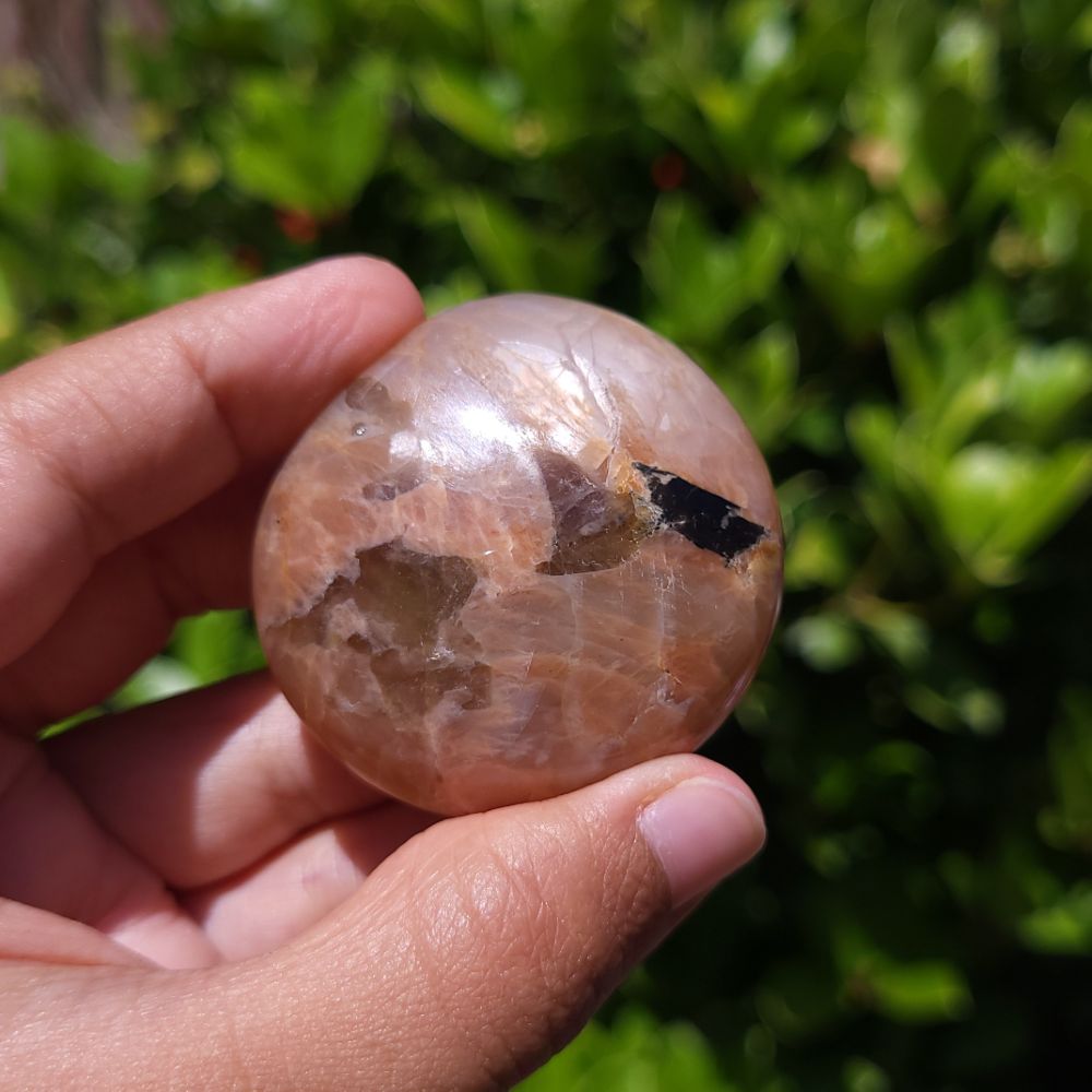 Hand holding a polished peach moonstone with smoky quartz inclusions with a blurred green background