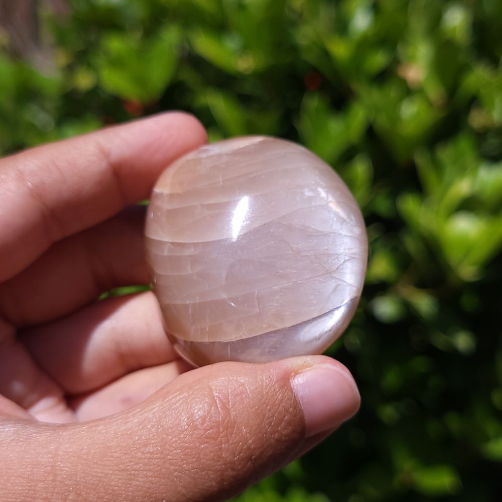 Hand holding a smooth, polished Peach Moonstone Palm Stone against a blurred green outdoor background