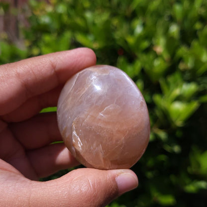 Hand holding a pink Peach Moonstone Palm Stone against a blurred green background