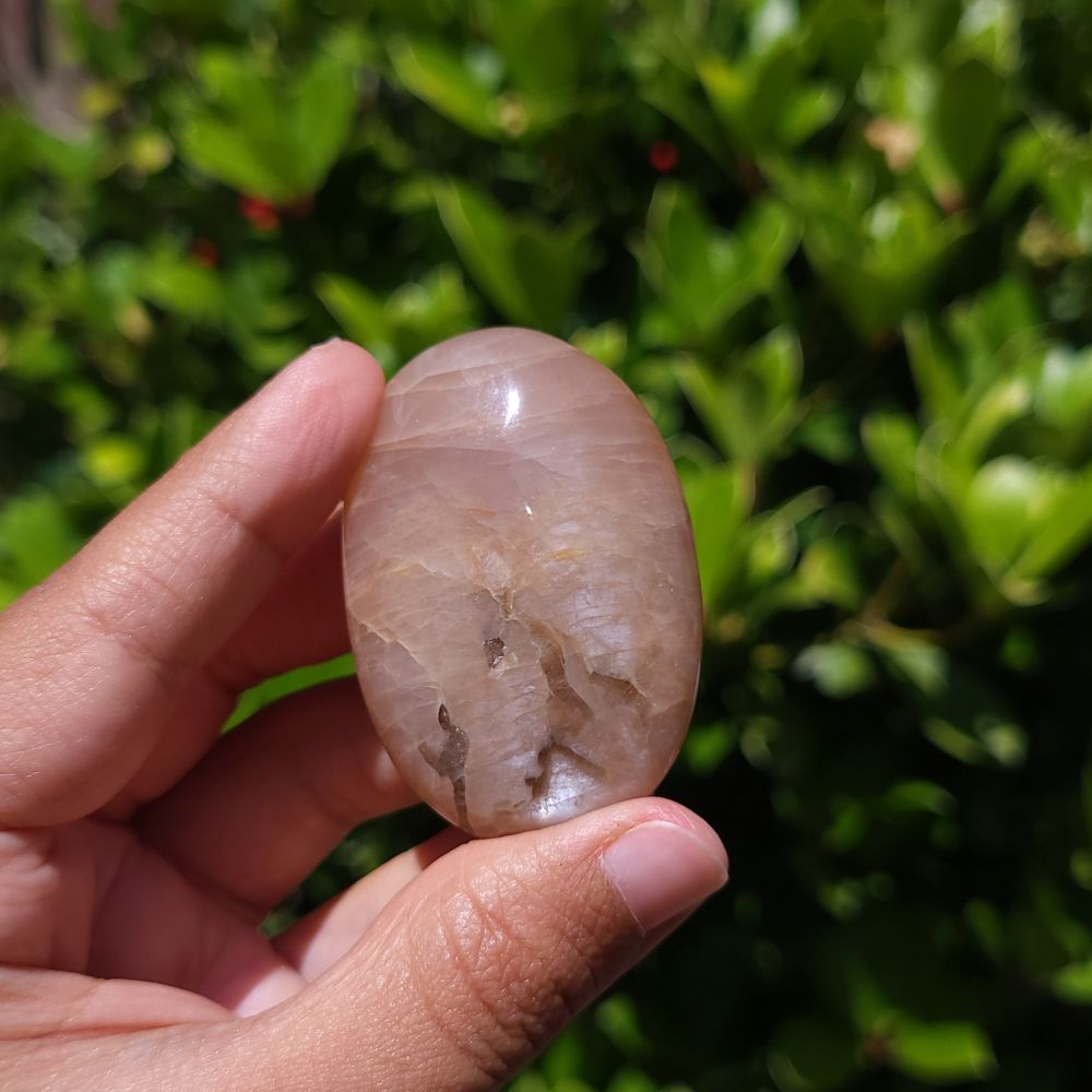 Hand holding a smooth Peach Moonstone Palm Stone with Smoky Quartz against a green leafy background
