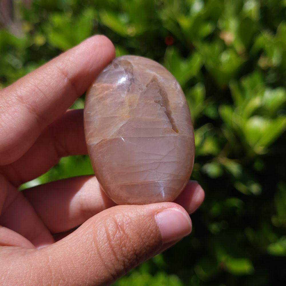 Hand holding a large Peach Moonstone Palm Stone with Smoky Quartz with a blurred green background