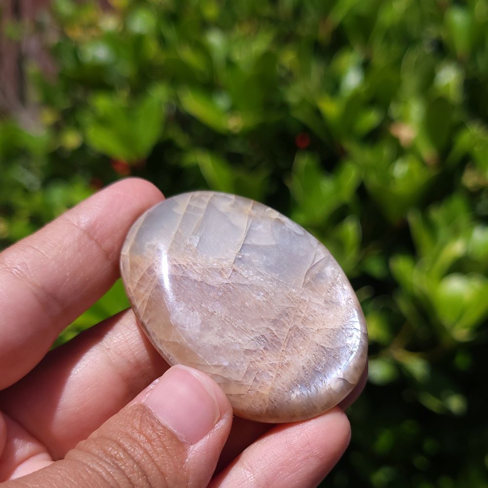 Hand holding a polished peach moonstone palm stone with a natural pattern against a blurred green background