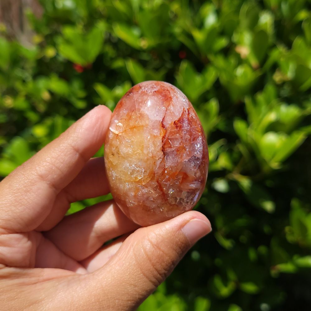 Hand holding a polished fire quartz palm stone against a green leafy background