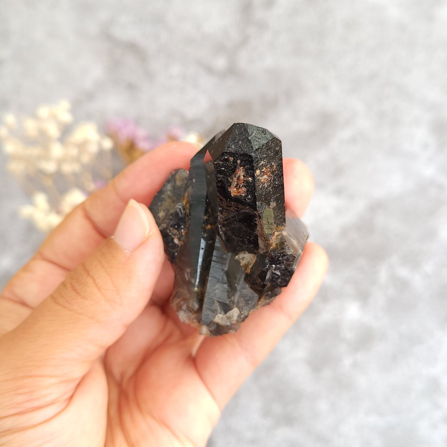 A hand holding a raw, faceted dark smoky quartz crystal cluster against a soft, blurred gray background with dried flowers.