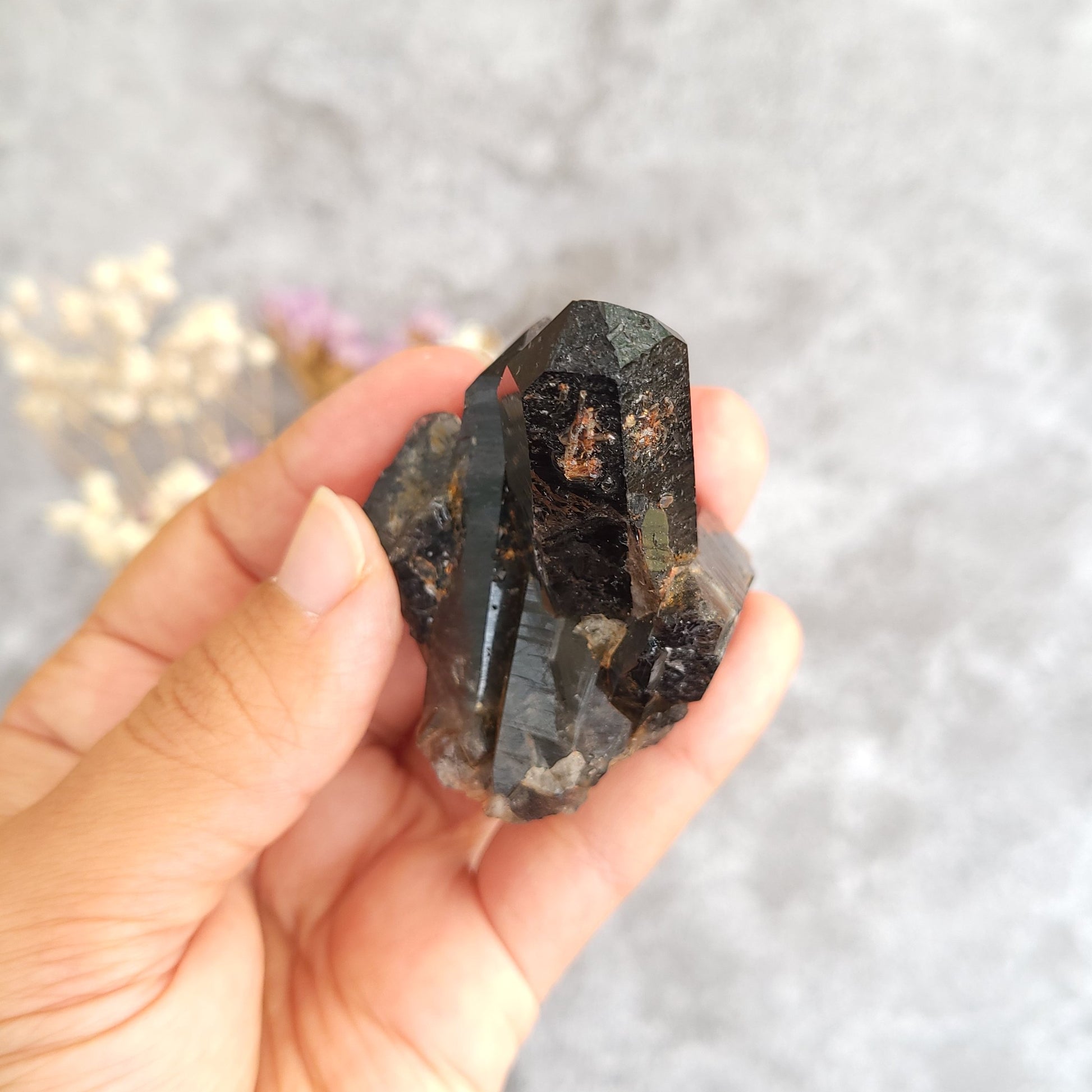 A hand holding a raw, faceted dark smoky quartz crystal cluster against a soft, blurred gray background with dried flowers.