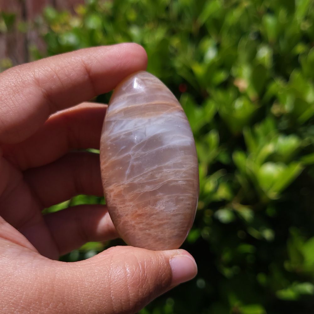 Hand holding a smooth peach moonstone palm stone against a blurred green background