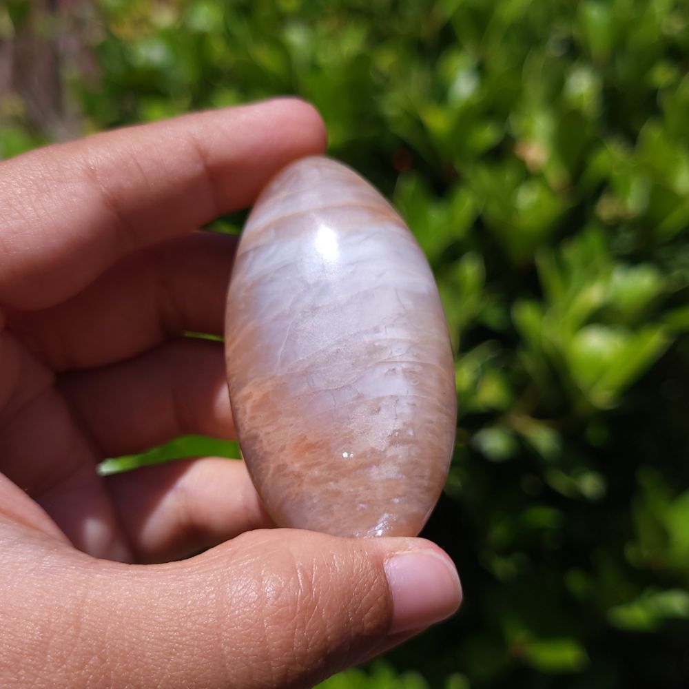Hand holding a smooth, oval-shaped peach moonstone palm stone with a blurred green background