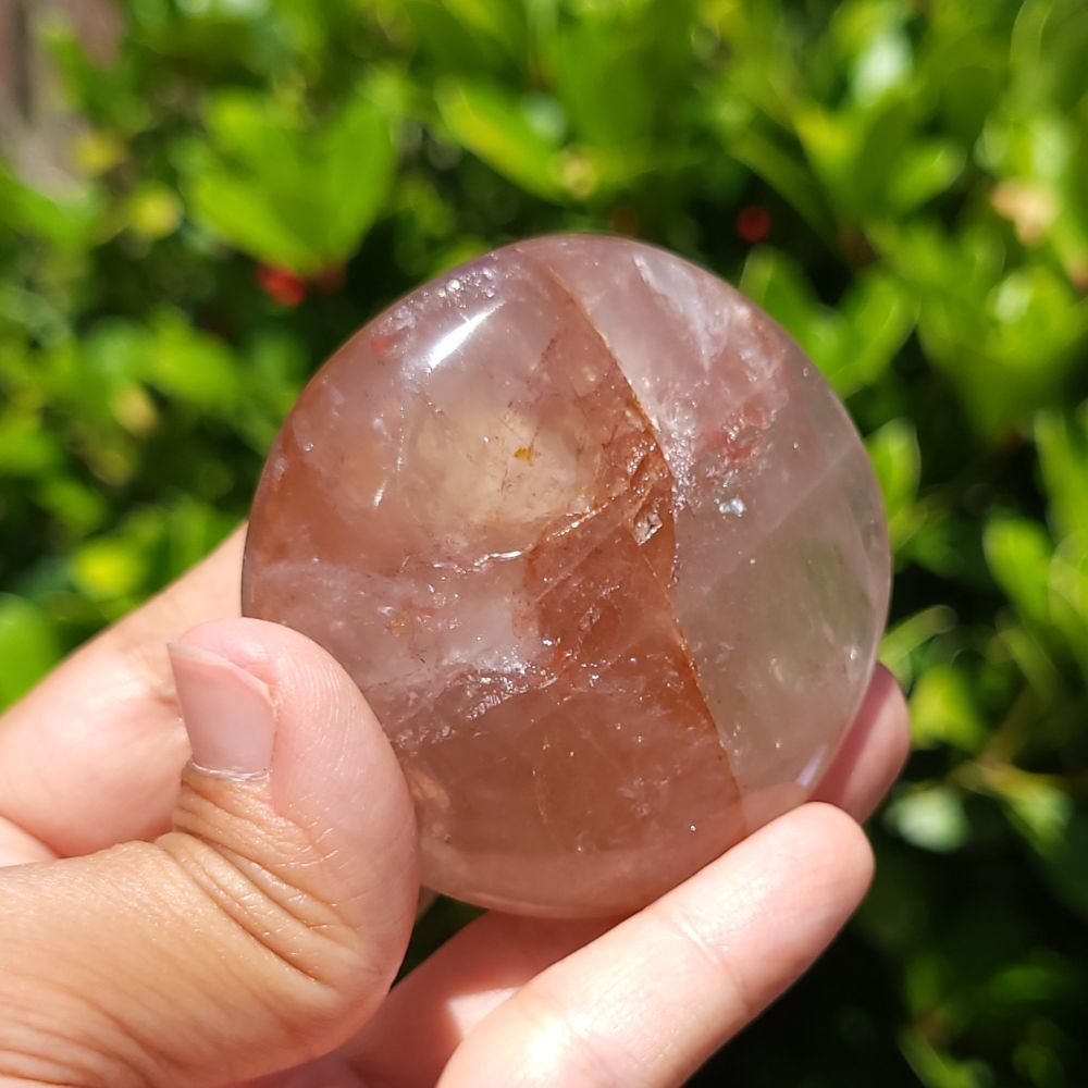 Hand holding a polished fire quartz palm stone with a green leafy background
