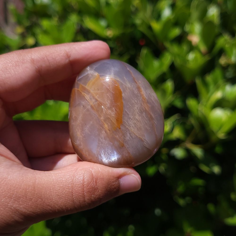 Hand holding a polished peach moonstone palm stone with a natural pattern against a green leafy background