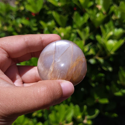 Hand holding a polished peach moonstone palm stone with a natural pattern against a green outdoor background