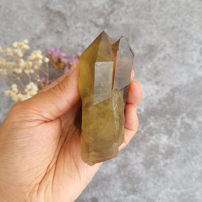 A hand holding a natural smoky quartz crystal cluster against a textured gray background with dried flowers in the soft focus.
