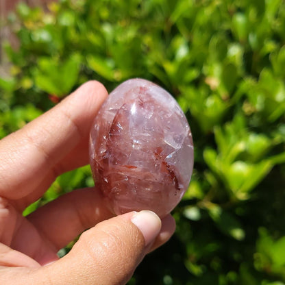 Hand holding a fire quartz palm stone with green foliage in the background