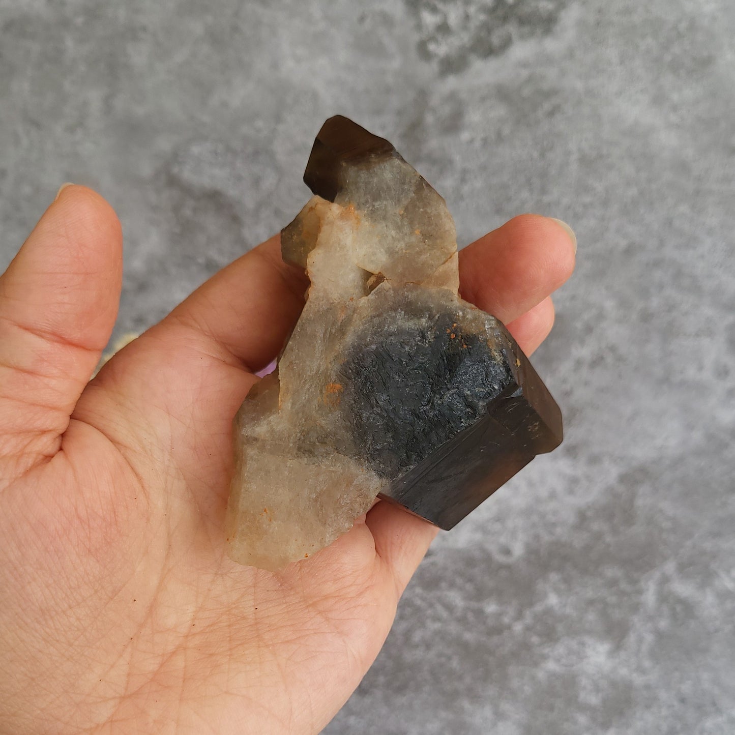 A hand holding a raw smoky quartz crystal cluster against a textured gray background.