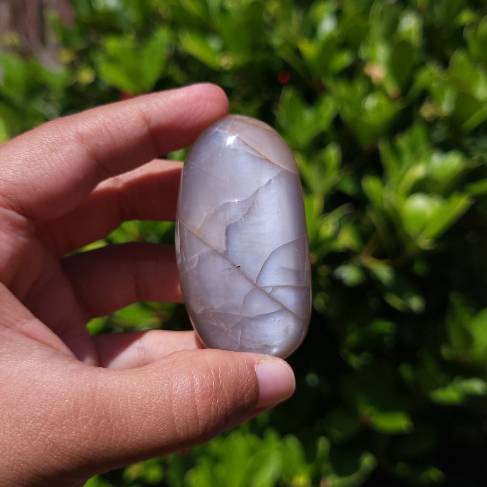Hand holding a polished peach moonstone palm stone with a natural pattern against a blurred green background