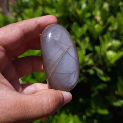 Hand holding a smooth, oval-shaped peach moonstone palm stone against a blurred green background