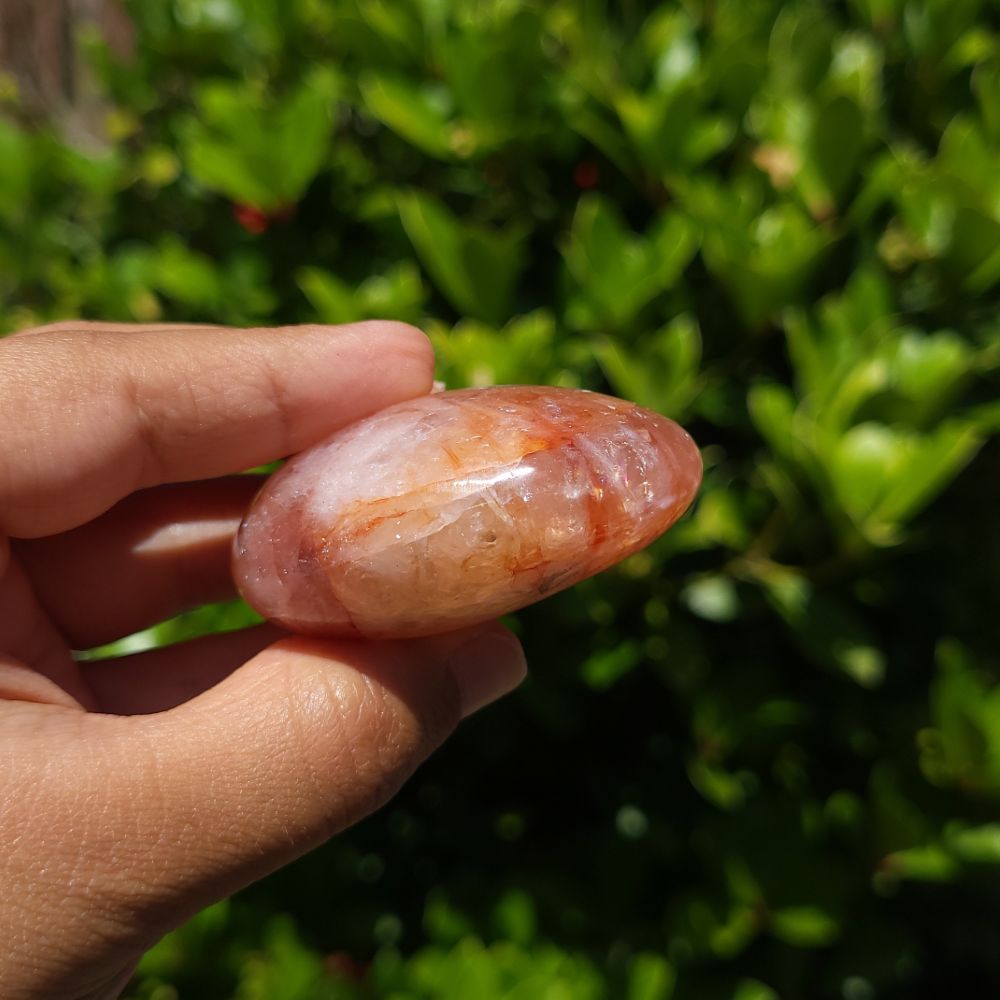 Hand holding a fire quartz palm stone with green foliage in the background