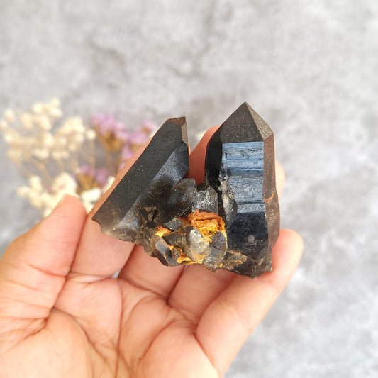 A hand holding a natural dark gray to black banded quartz crystal cluster with metallic gold and orange inclusions, set against a soft gray textured background with blurred dried flowers.