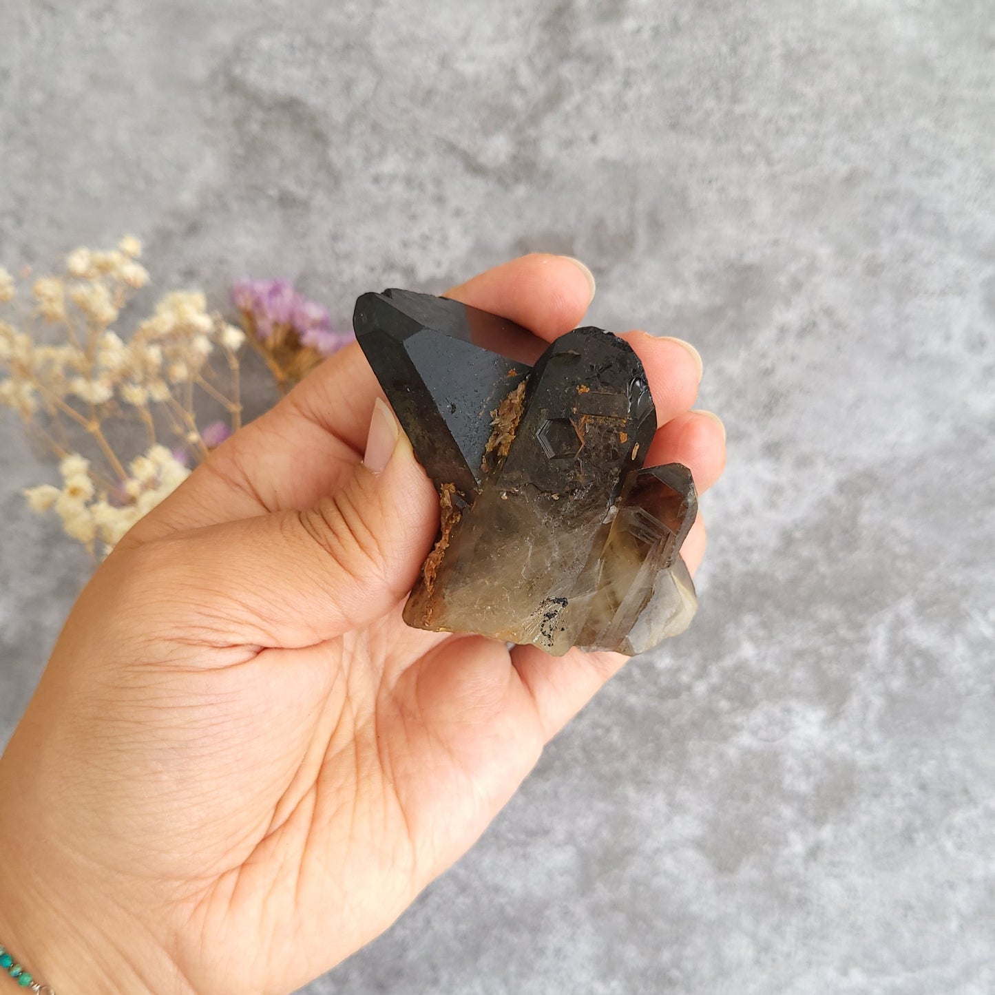 A hand holding a smoky brown and translucent crystal cluster against a textured gray background with dried flowers.