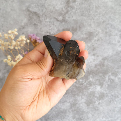 A hand holding a smoky brown and translucent crystal cluster against a textured gray background with dried flowers.