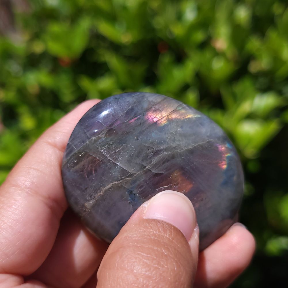 Hand holding a purple flash labradorite palm stone with a blurred green background