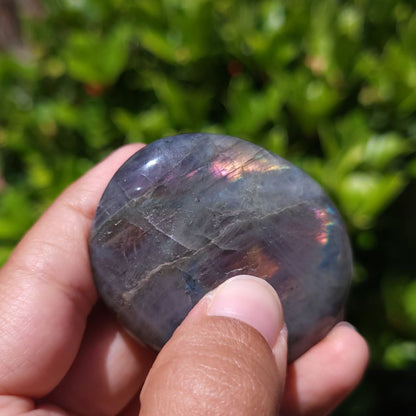 Hand holding a purple flash labradorite palm stone with a blurred green background