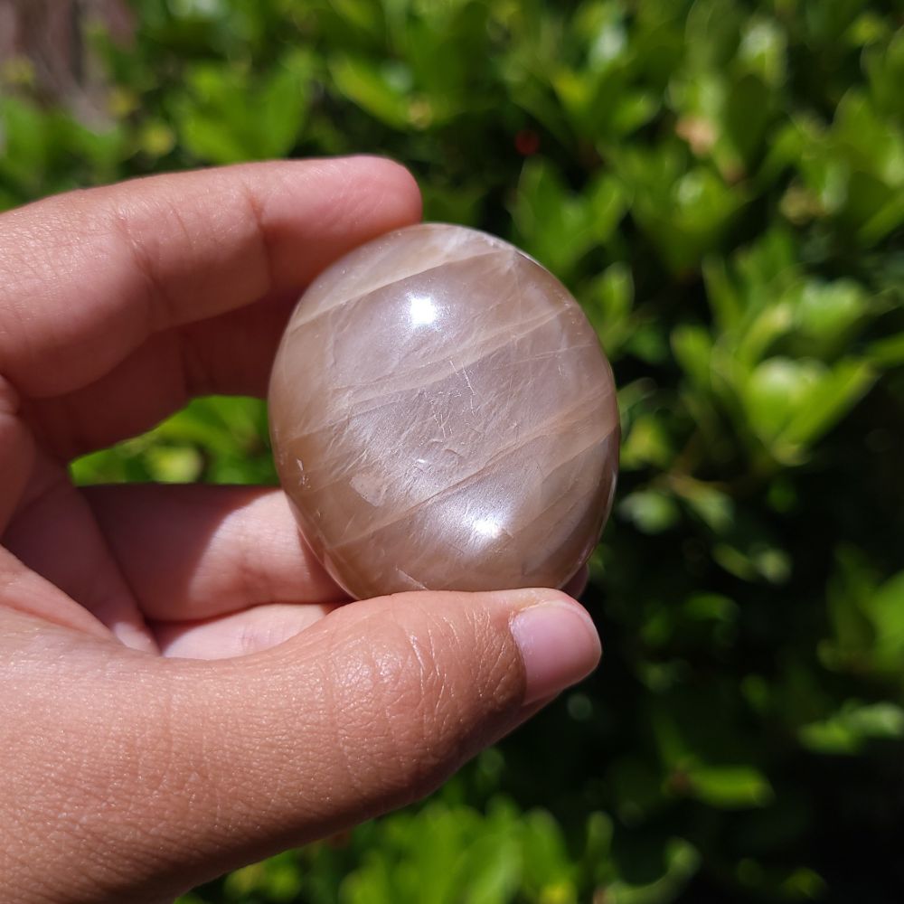 Hand holding a smooth, peach moonstone palm stone against a green outdoor background