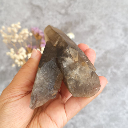 A hand holding two rough, translucent smoky quartz crystal points against a soft gray textured background with blurred dried flowers.