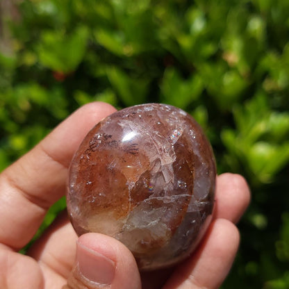 Hand holding a Fire Quartz palm stone against a green leafy background