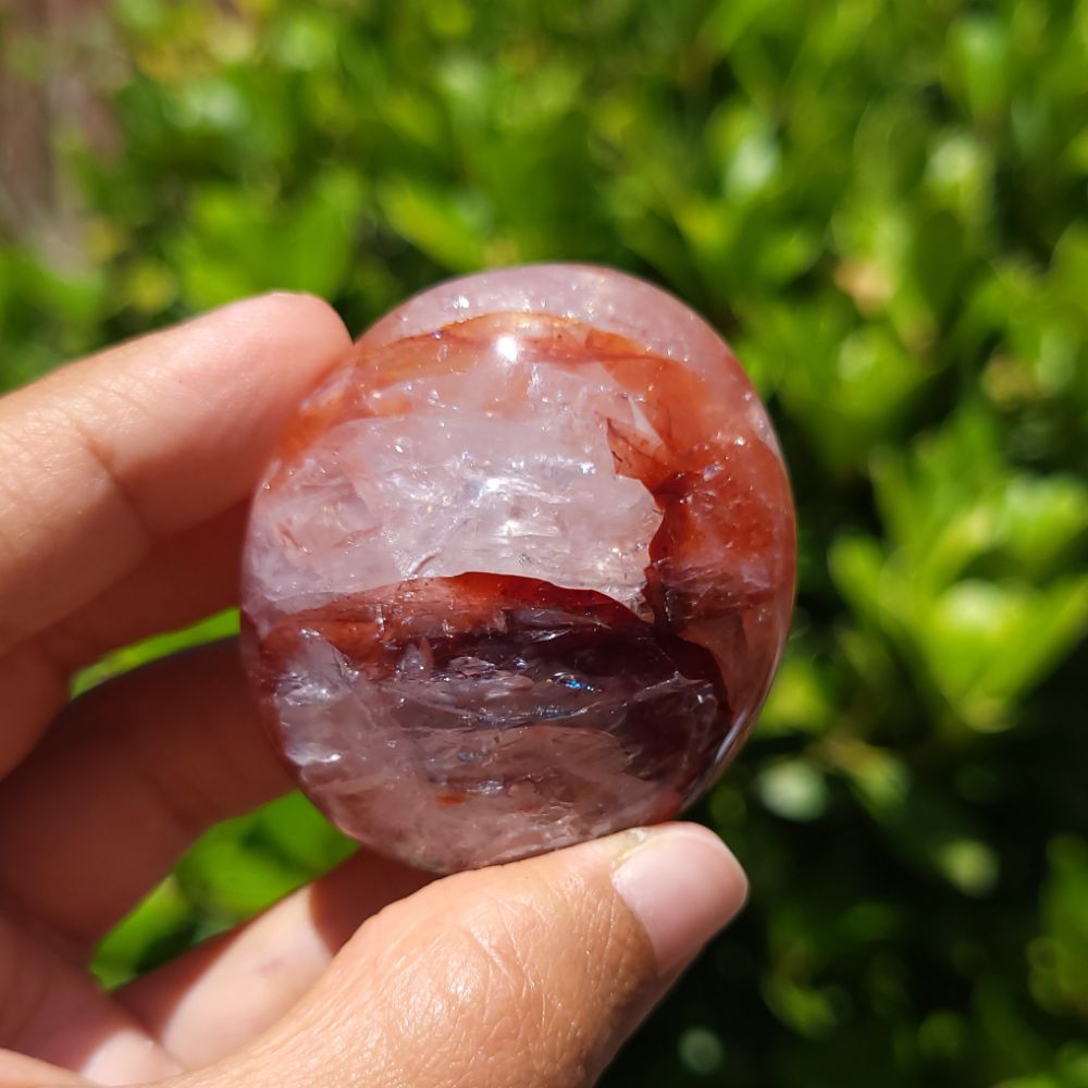 Hand holding a Hematoid Quartz palm stone crystal against a green leafy background