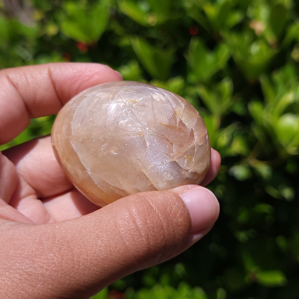 Hand holding a smooth, polished Peach Moonstone palm stone against a green outdoor background