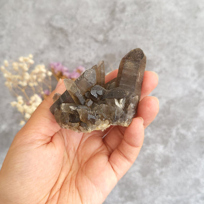 A hand holding a natural smoky quartz crystal cluster against a textured gray background with blurred dried flowers.